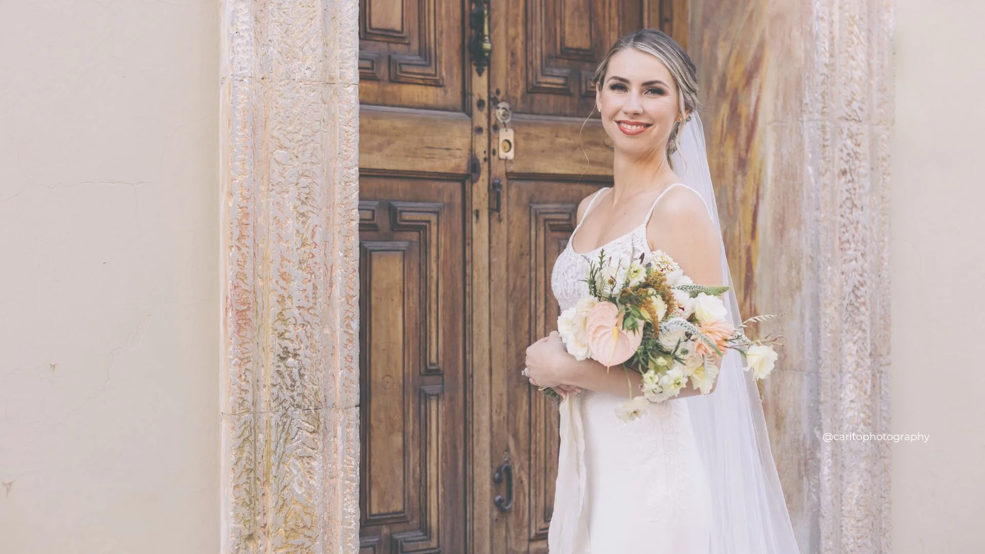 Novia con maquillaje luminoso sosteniendo un ramo pastel frente a puertas de madera tallada