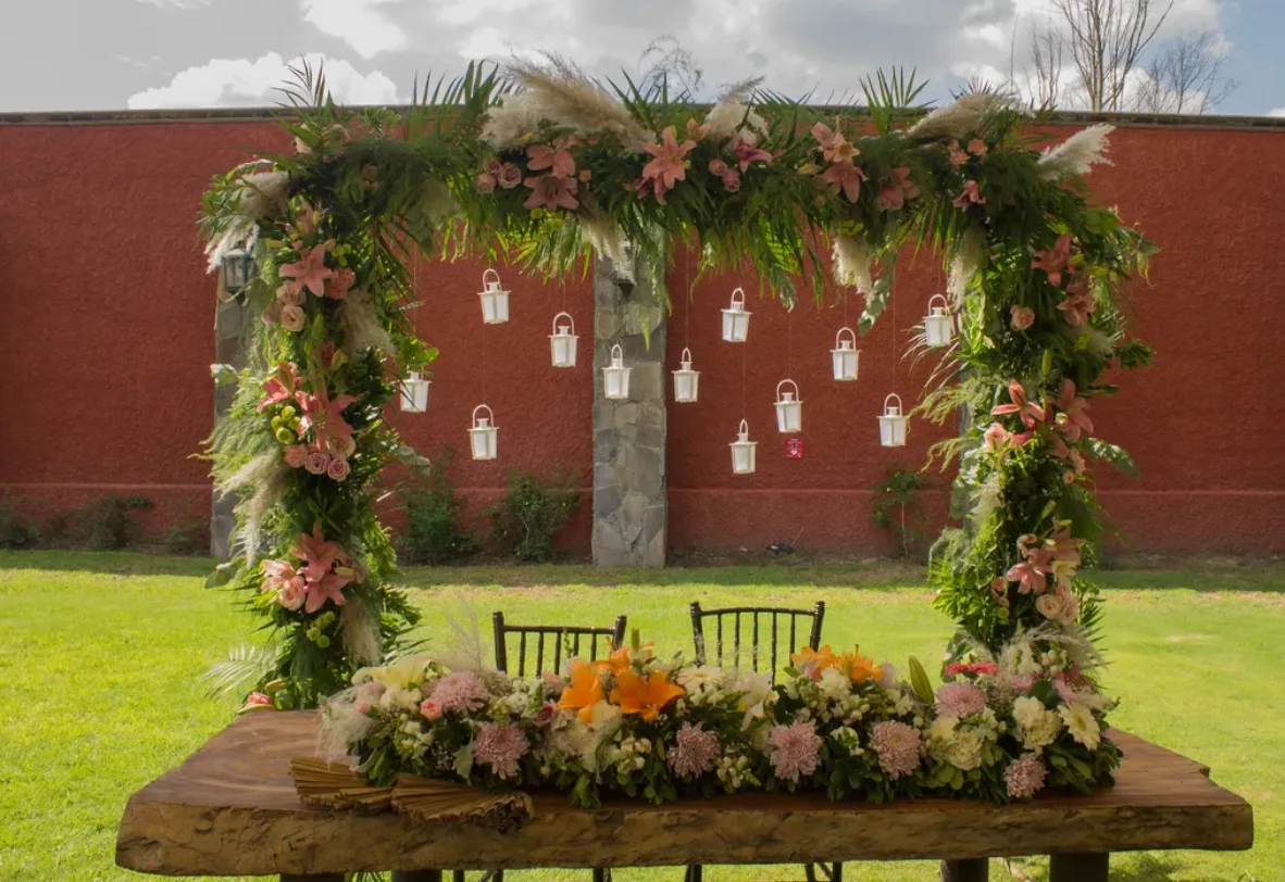 Estructura de boda en jardín cubierta de follaje, flores rosas y faroles colgantes de Florería Florena