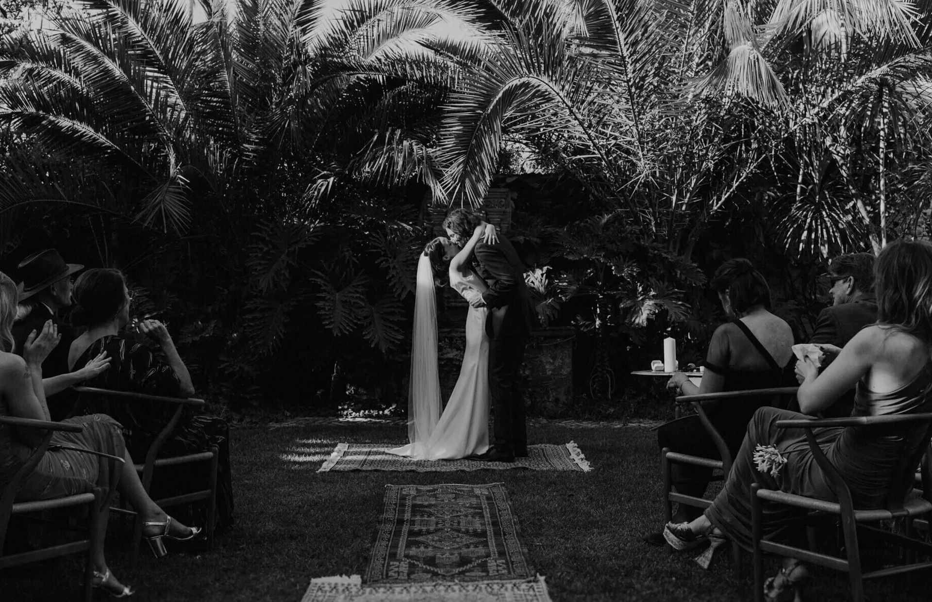 Fotografía documental en blanco y negro de una pareja besándose durante una ceremonia en jardín