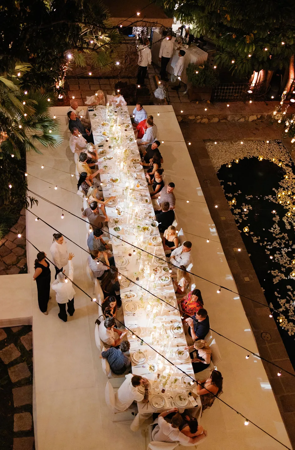 Vista aérea de una mesa de banquete iluminada con velas junto a un estanque cubierto de pétalos