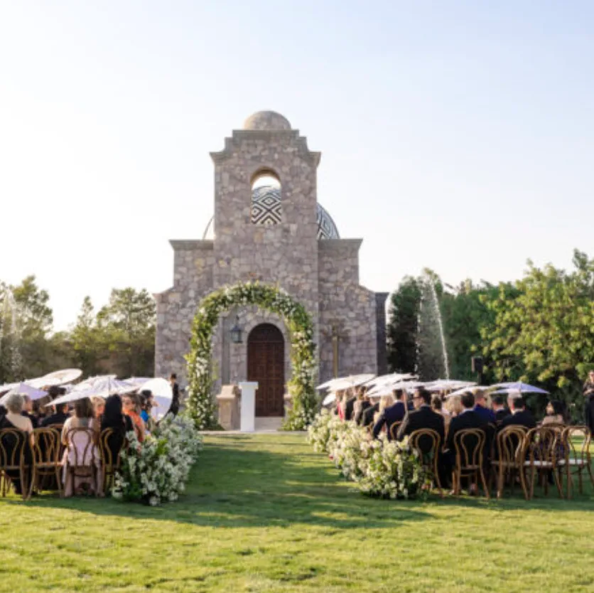 Ceremonia de boda al aire libre frente a una capilla de piedra con pasillo enmarcado por flores