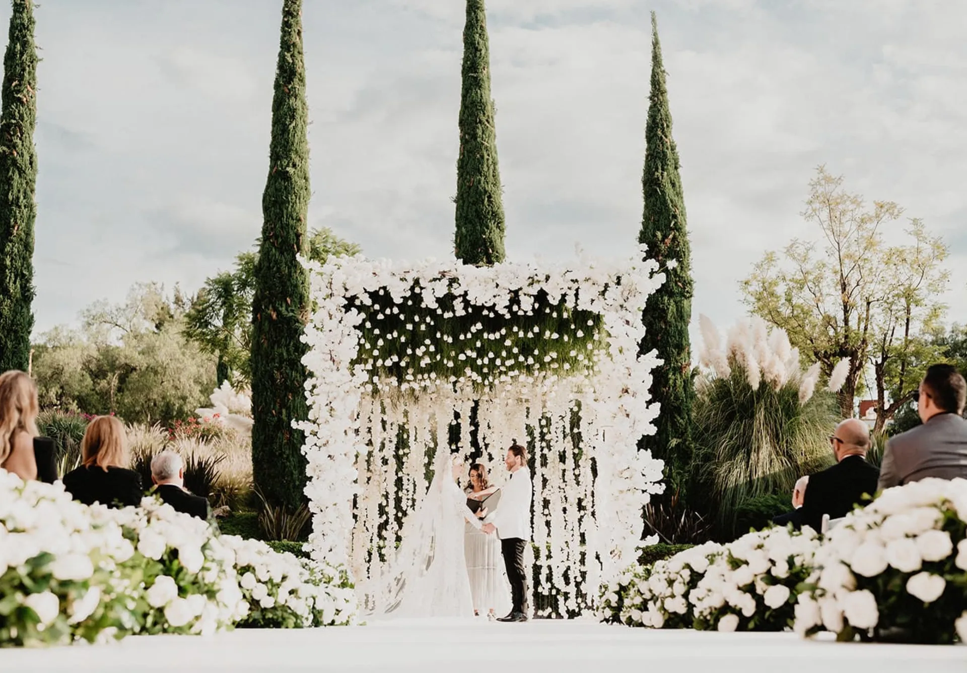 Ceremonia de boda al aire libre enmarcada por flores blancas colgantes y altos cipreses