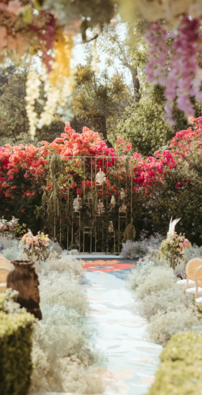 Pasillo de ceremonia en jardín con flores blancas etéreas, detalles coloridos y flores suspendidas