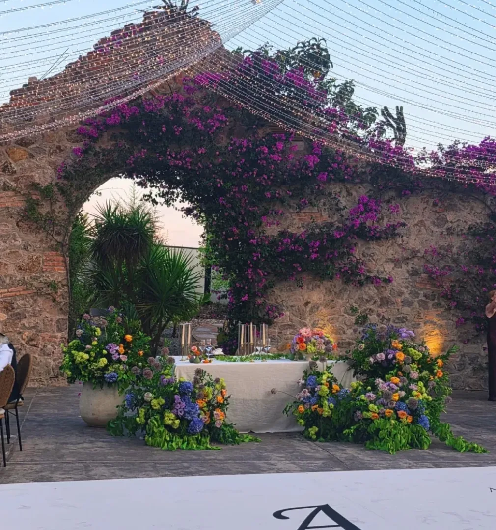 Mesa de novios con flores coloridas enmarcada por arcos de piedra y buganvilias en Hacienda Agualuna